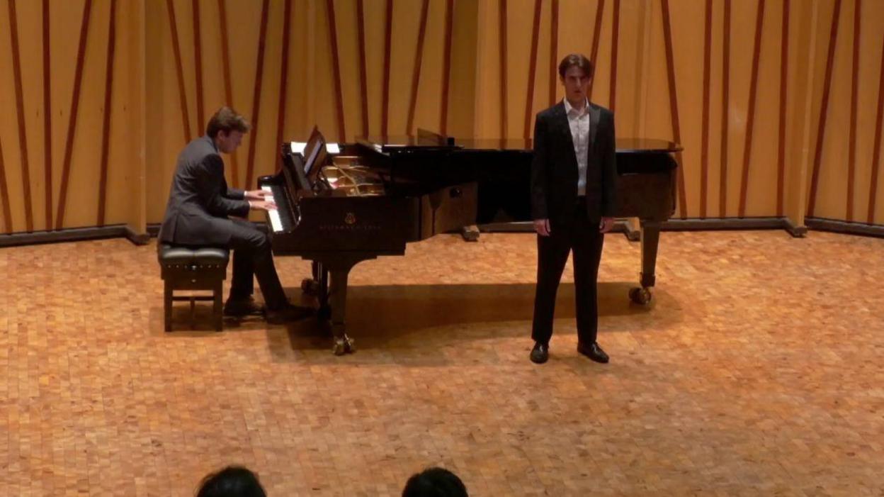 Alex Harpum sings in front of an audience with his feet shoulder width apart. He is wearing a black dinner jack, black trousers and white, open-collared shirt. A pianist accompanies his singing in the background of the still picture, taken from a video of the performance. The silhouettes of the heads of members of the audience can be seen in the foreground of the image.