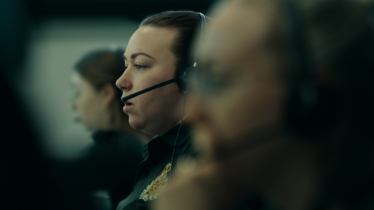 Three women are sat down looking to the left of the picture. Everywhere is blurred apart from the woman in the middle who has a headset on.