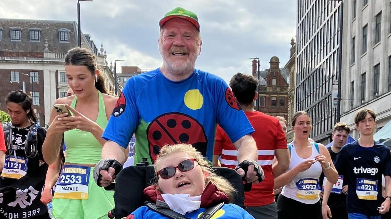 A man with a green hat, and blue T shirt with ladybirds on smiles at the camera ahead of a running race. He is pushing a wheelchair with a young woman in it. She has long blonde hair and is wearing red sunglasses.