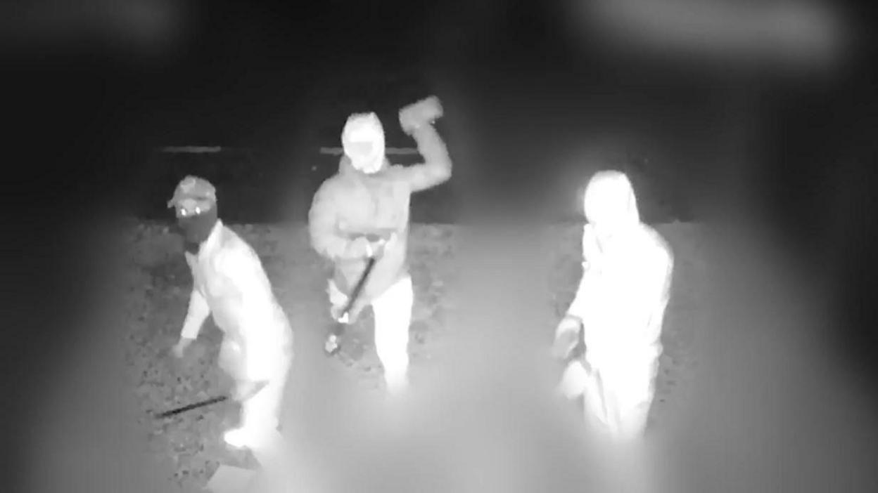 A black and white image showing a screenshot from CCTV footage. There are three individuals with their faces covered, wearing tracksuits and coats. They are carrying large machetes and throwing bricks through the window of the property. 