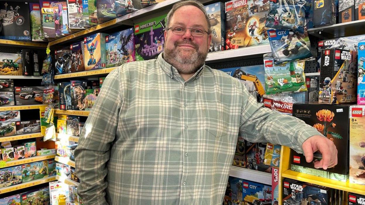 Matt Booker leaning on shelves inside his shop with toys and their boxes on them. He is smiling at the camera and is wearing glasses