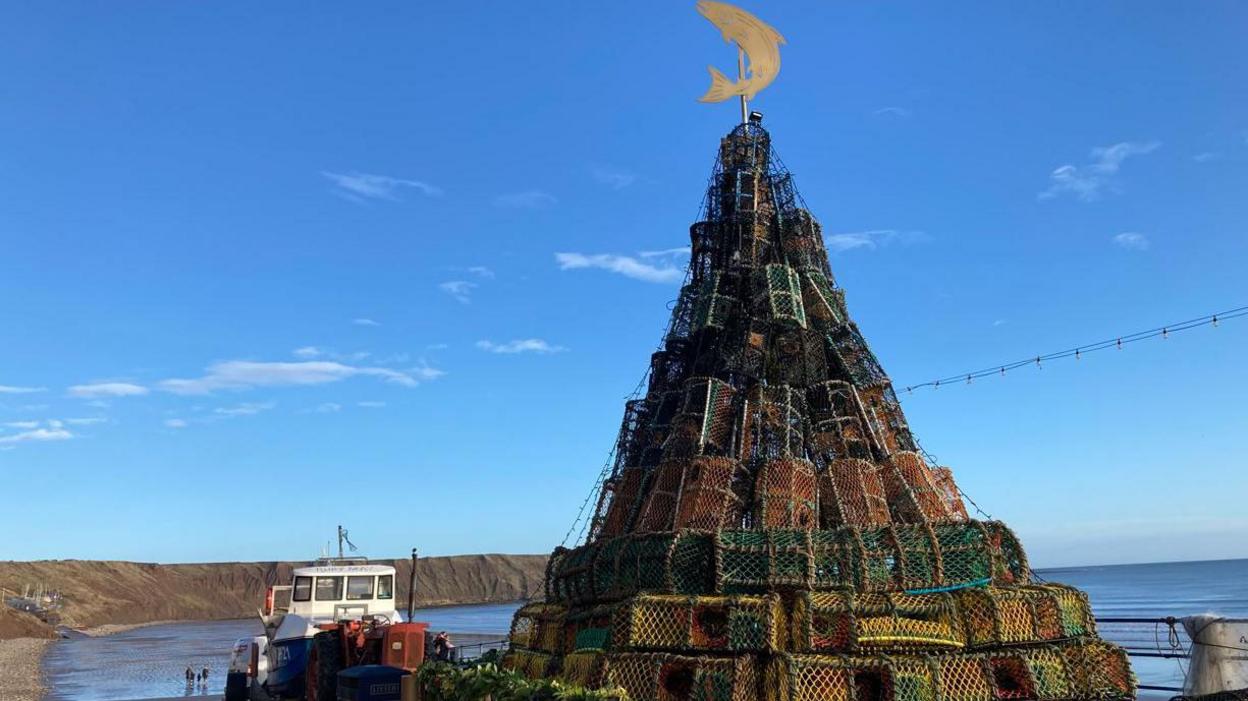 A large Christmas tree built from lobster pots, with a view of the North Yorkshire coast in the background. It's a sunny day, with people walking on the beach. A large fishing boat is also in the background. 