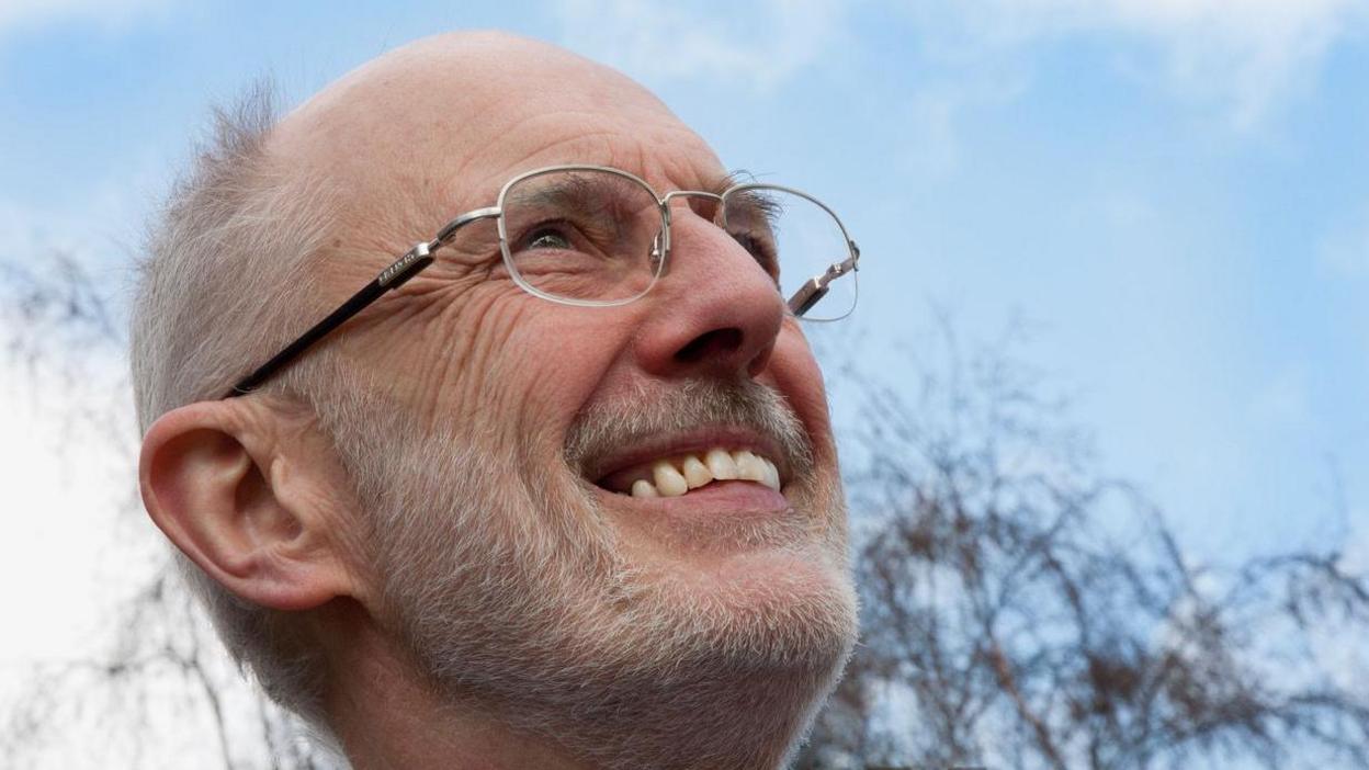 A close-up of Paul Andersen, who has short grey hair and beard and wears silver-rimmed glasses. He is smiling and looking up at a blue sky.