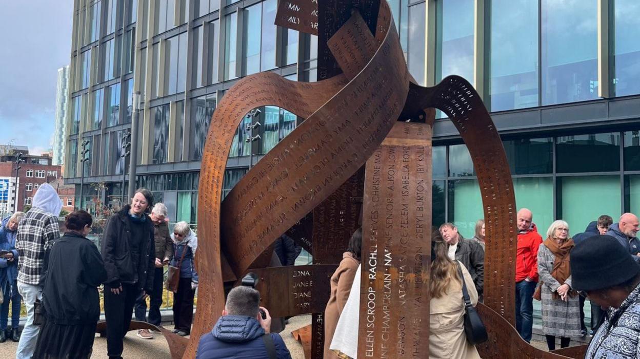 People stand around a bronze statue of ribbons with names imprinted on them