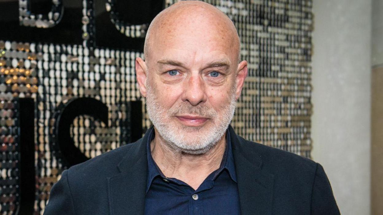 Brian Eno in a shirt and suit standing in front of a silvery background in 2015