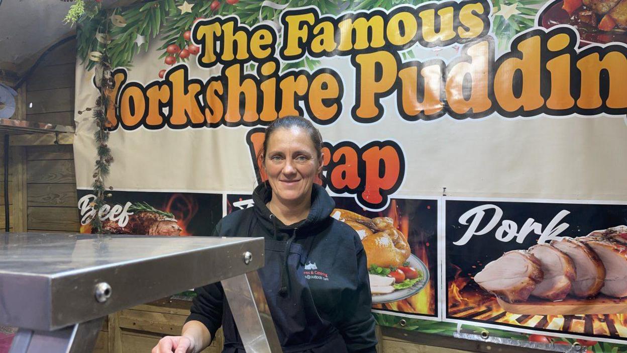 A lady in a blue top smiling to camera in front of a banner saying 'The Yorkshire Pudding Wrap' and standing behind a food counter