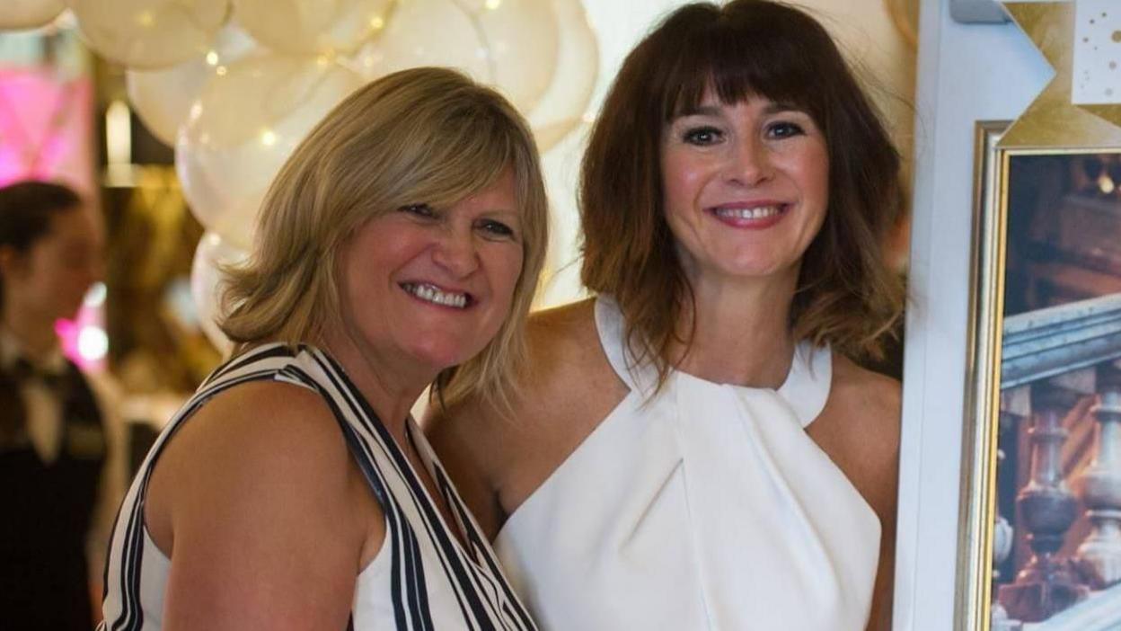 Smiling Angela MacVicar and Lindsay MacCallum wearing party dresses posing next to a photograph of Johanna MacVicar with helium-filled balloons behind them