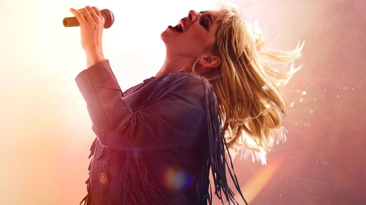 Dawn Sievwright performing as Rose-Lynn Harlan, wearing a tasselled leather jacket and large hooped earrings. She is holding a microphone and has her head held back with her blonde hair flying behind her.