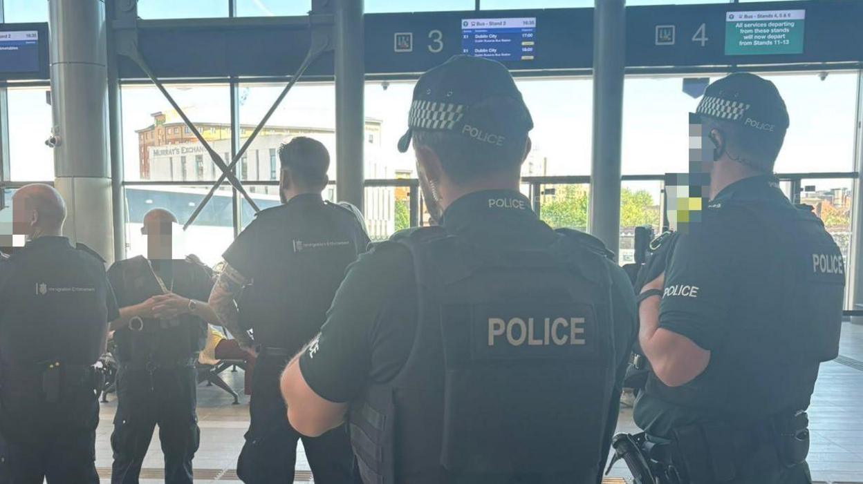 The picture shows from behind police officers wearing dark green uniforms, baseball caps and flak jackets. In from of them are three members of a Home Office enforcement team, also wearing dark uniforms. The picture is taken inside a bus station