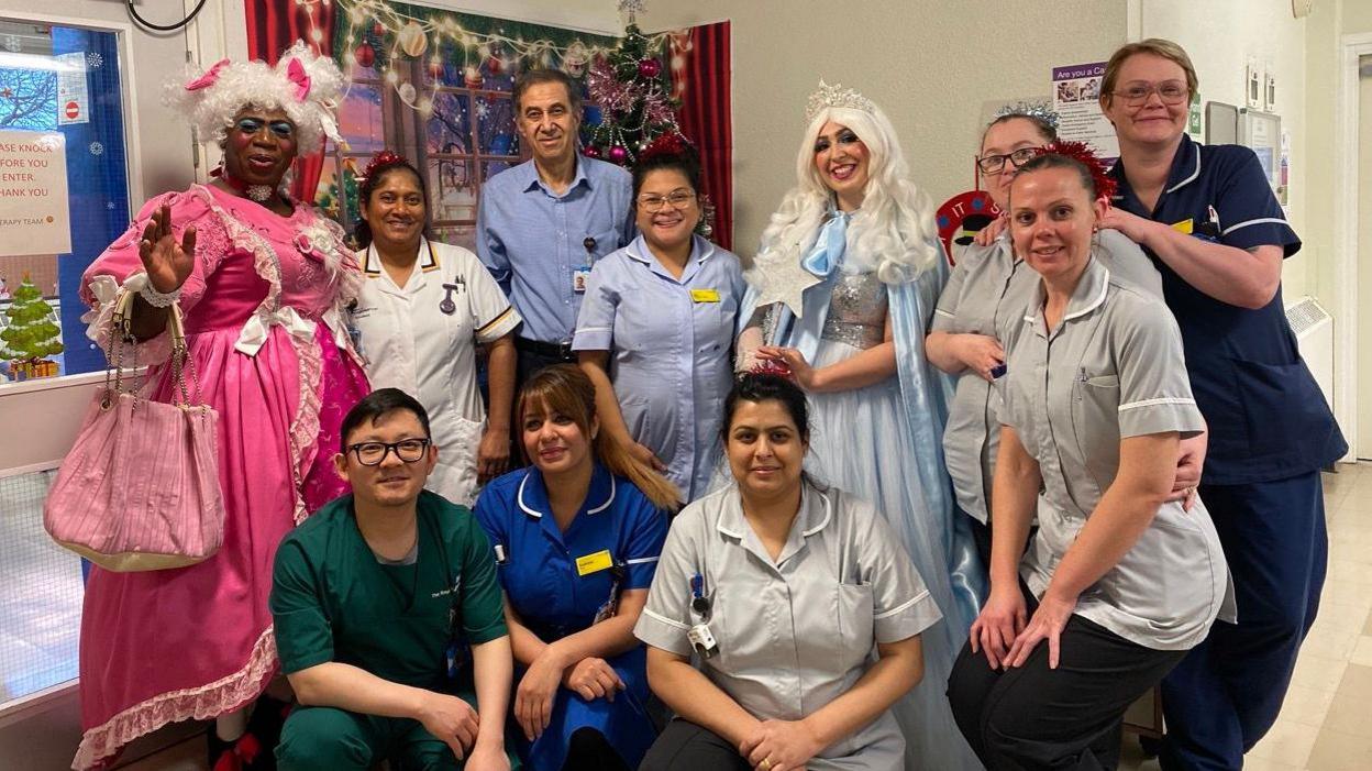 A group of hospital staff wearing uniforms are standing next to the two actors who are wearing their costumes. They are standing in the hospital ward in front of a Christmas tree.