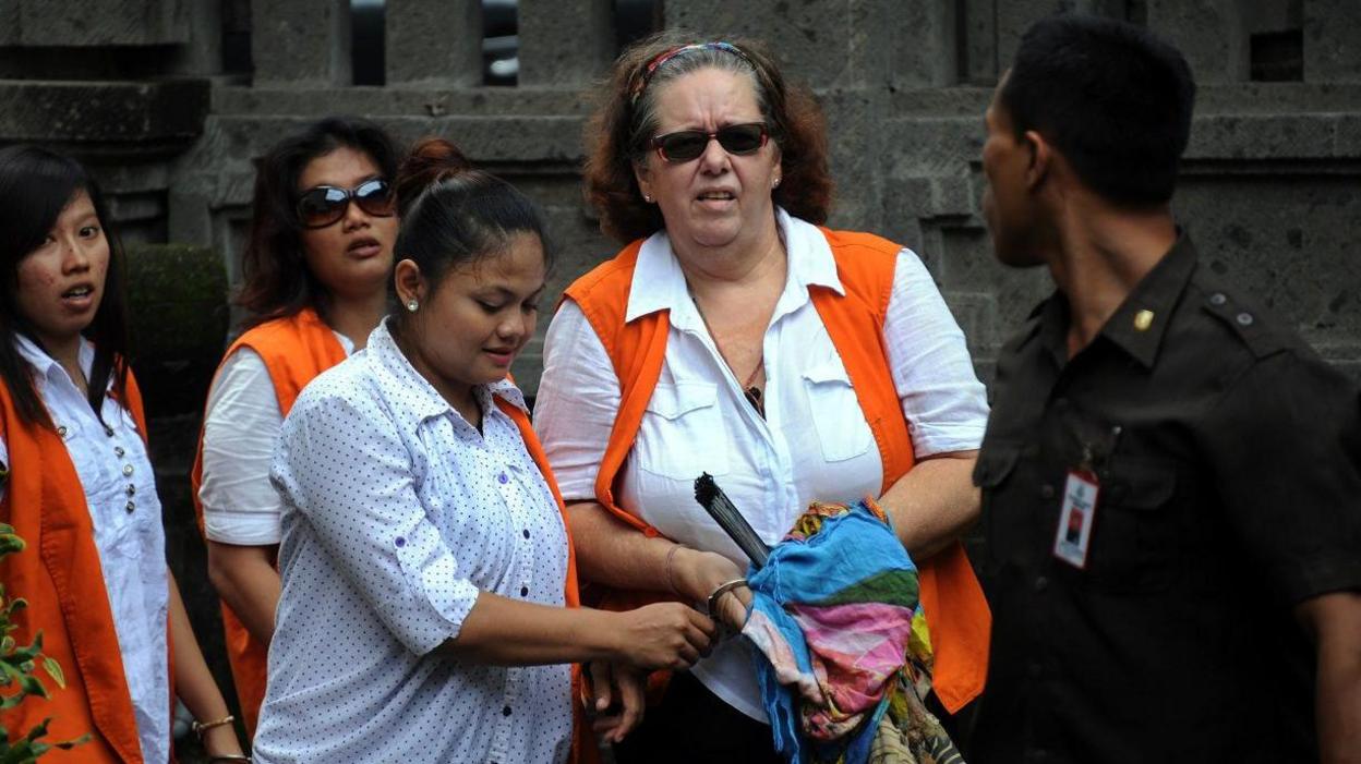 Lindsay Sandiford, who has short curly hair, walking alongside Indonesian officials. One is dressed in a green uniform and the others are wearing white blouses with orange tops.