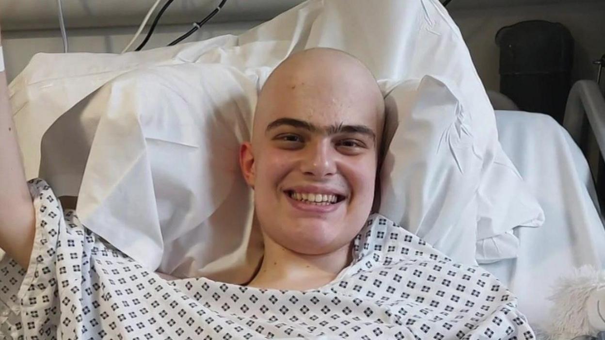 Harry, who wears a hospital gown, smiles from a hospital bed.