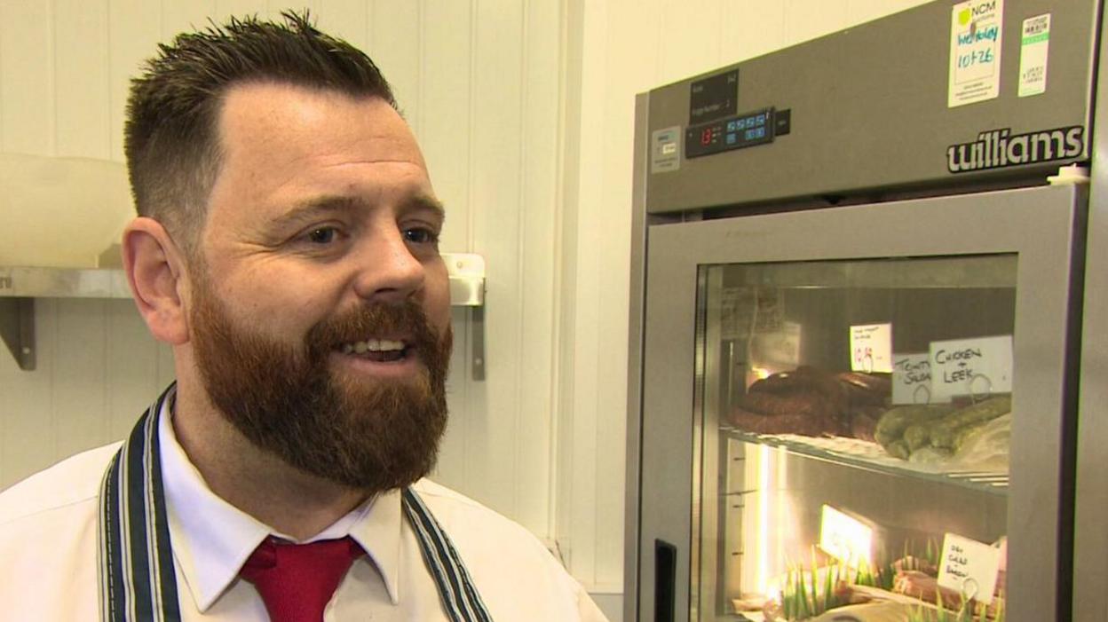 A butcher stands in his shop in front of a fridge full of meat. He has short, spiky hair and a full brown beard and is wearing a white shirt, red tie and blue and white striped apron.