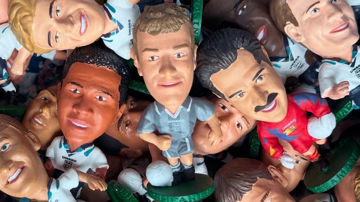 A bundle of plastic football figures on round green bases with large heads. They are England players wearing the white Euro 96 kit while Alan Shearer is in a grey strip and David Seaman in a red one