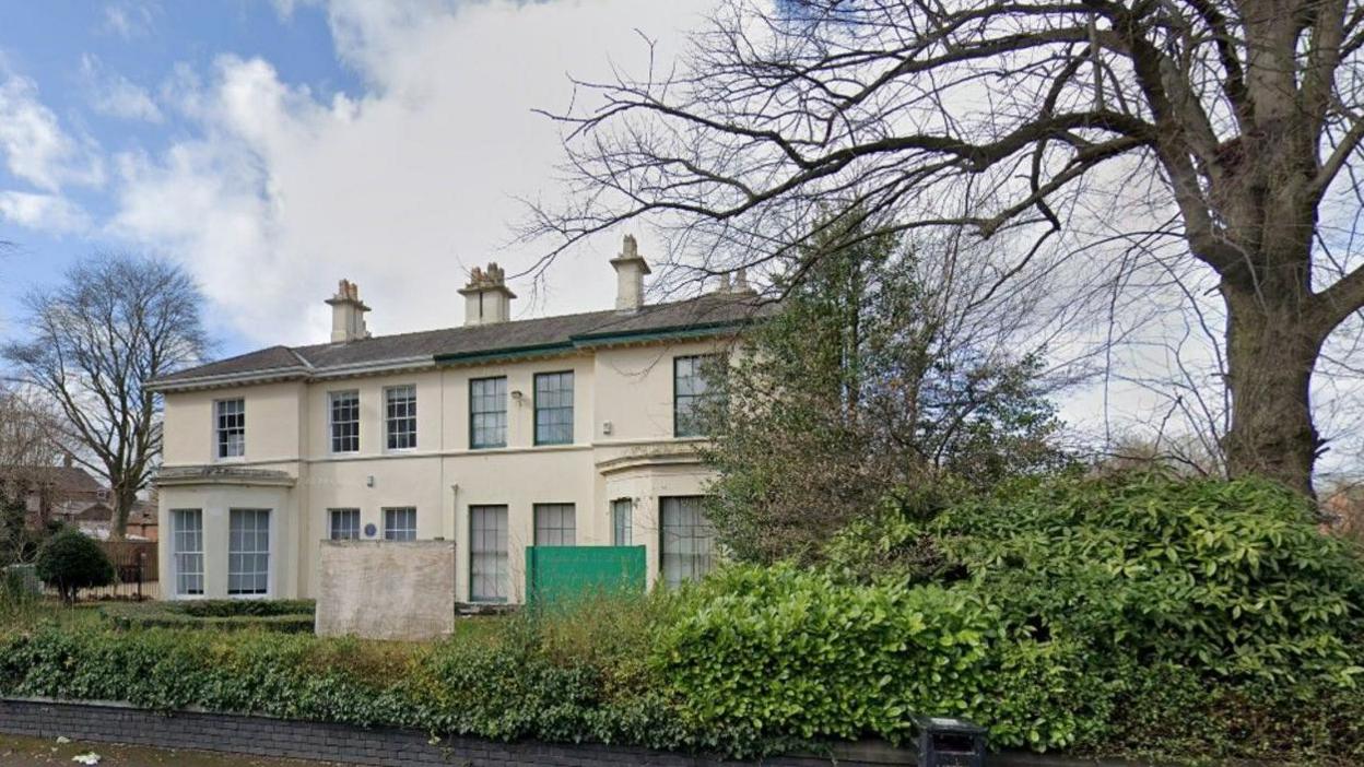 A light-coloured two-storey building has a tree to the left of it on the photo. A hedge is above a small brick wall in the foreground.