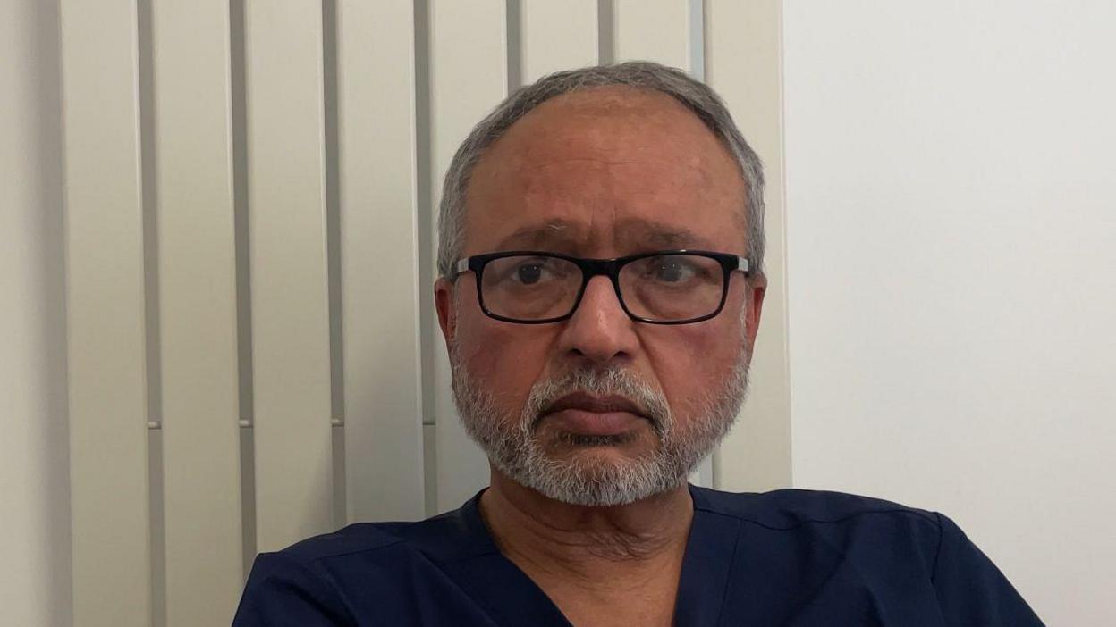 A grey haired man with grey beared and black glasses looks at the camera. He is wearing medical scrubs.