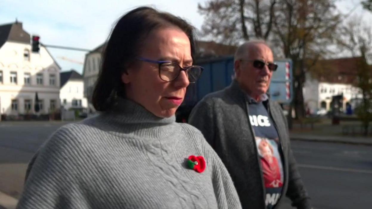 Natasha Walker walks in a street beside her father Richard Lee. She had brown, shoulder-length hair and glasses and wears a poppy badge on a light-grey sweater. He has grey hair and wears sunglasses and a sweatshirt with a picture of Katrice in a red dress.