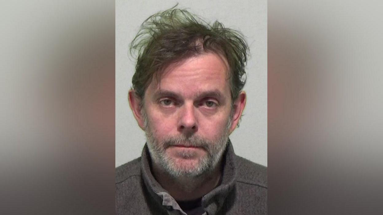 Mugshot of William Henderson who messily ruffled dark hair and a grey beard.