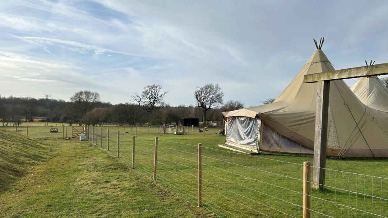 A pleasant day on farm land. There is a tee-pee and some animals in the background. There are clouds in the sky and some trees can be seen in the background. 