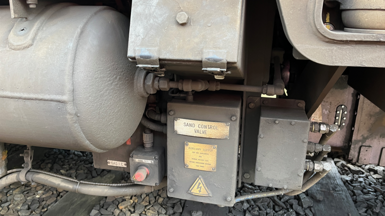 A close-up of a grey metal box underneath a train with SAND CONTROL VALVE written on a gold plaque.