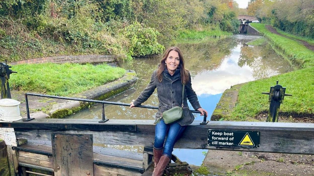 A woman sits on a wooden canal lock with the waterway visible behind her. She has long dark hair, and is wearing a grey jacket, blue jeams and brown leather boots.