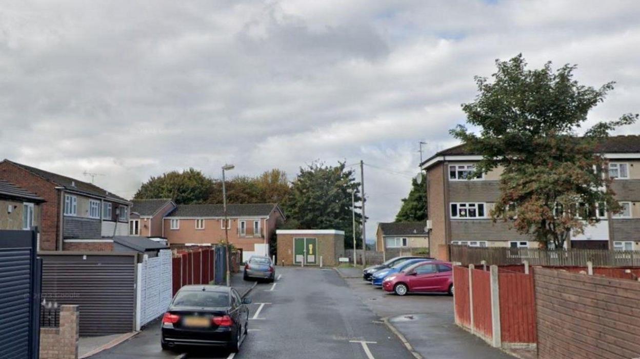 A generic image of a residential street on a cloudy day. There are houses on the left and there is a three-storey block on the right. There are cars parked on both sides of the street.
