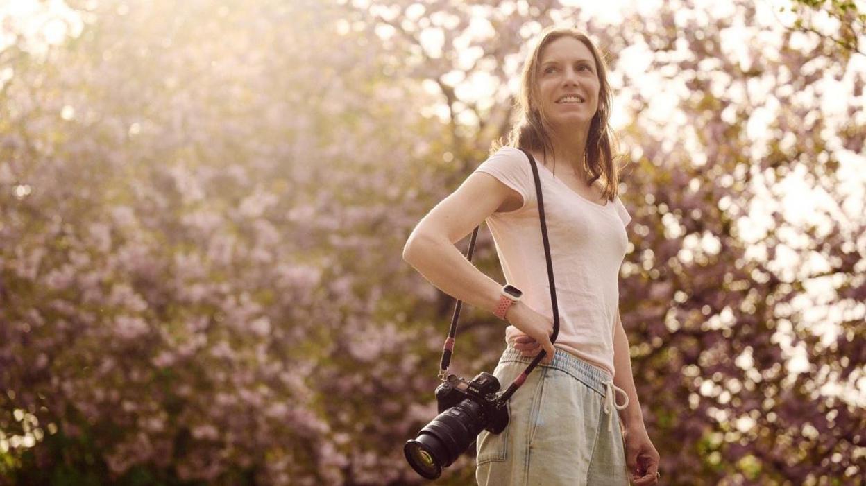 Bethany Clarke, a woman with long brown hair wearing a pink t shirt and trousers, holds her camera as she stands beneath a cherry blossom tree