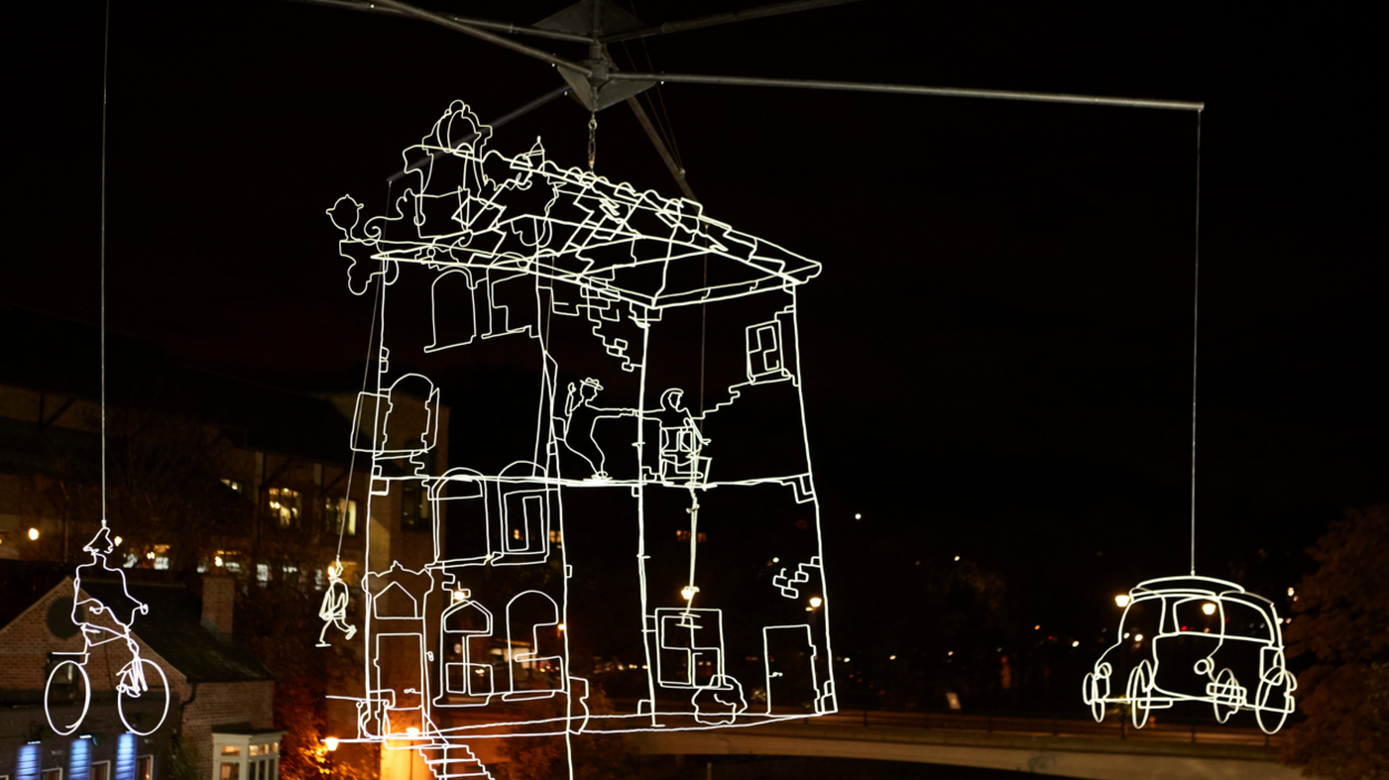 Drawn in Light is an installation made of steel, hand-formed into a typical Amsterdam-style canal building. It is surrounded by a man on a bicycle and a car. The artwork shines in bright yellow. 