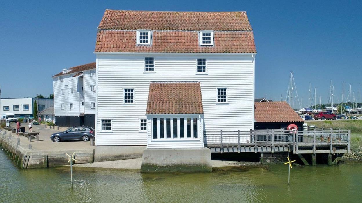 Tide mill on the River Deben. It is a large white wooden-clad four-storey building sitting on the waterfront. The masts of boats can be seen to the right of the mill