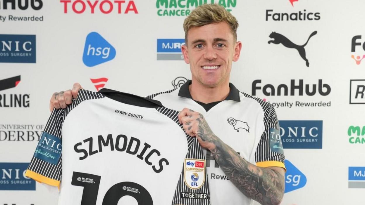 New Derby forward Sammie Szmodics holds up a Rams shirt with his surname and number
