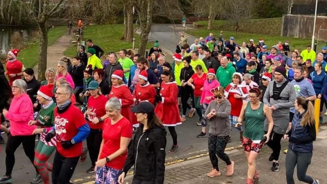 Dozens of people, some in festive costumes, running. Trees and grass are in the background.