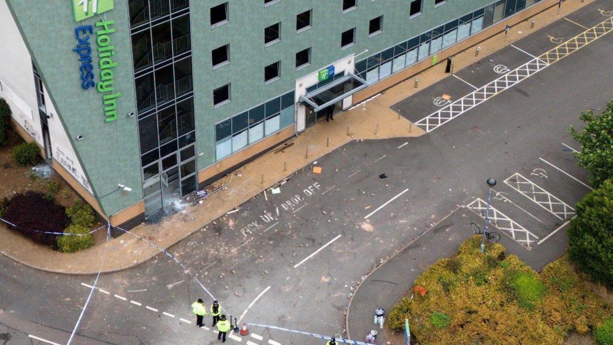 An aerial view of a hotel building with a blue fascia, along the left side is a green and blue sign that says Holiday Inn Express. In front of the hotel is a road and car park that has been emptied, but shows signs of disorder as there is broken glass and rubble visible. A police cordon is in place towards the bottom of the image with three officers standing next to it