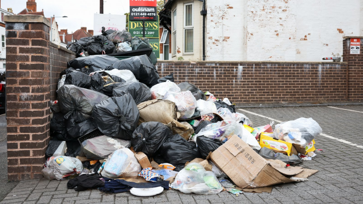 A very large pile of bin bags stacked against a brick wall. Houses can be seen behind the brick wall.