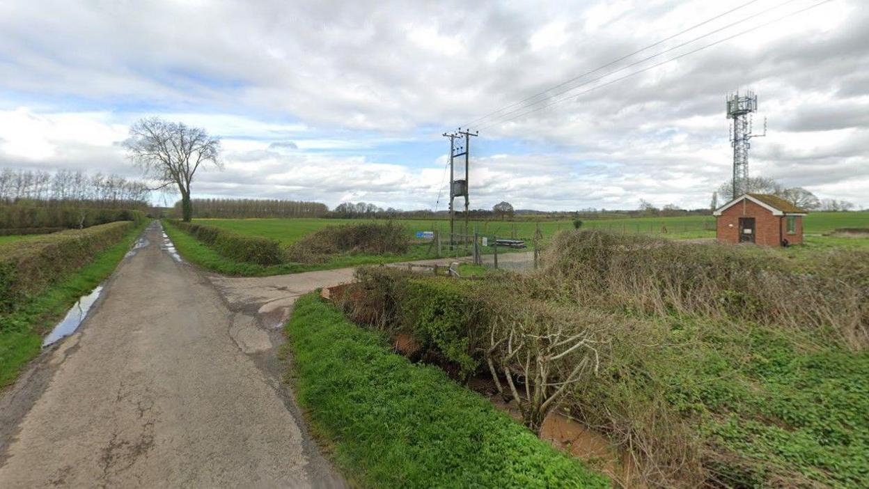 A track runs between two sets of fields with a small brick-built building off to the right behind a set of hedgerows