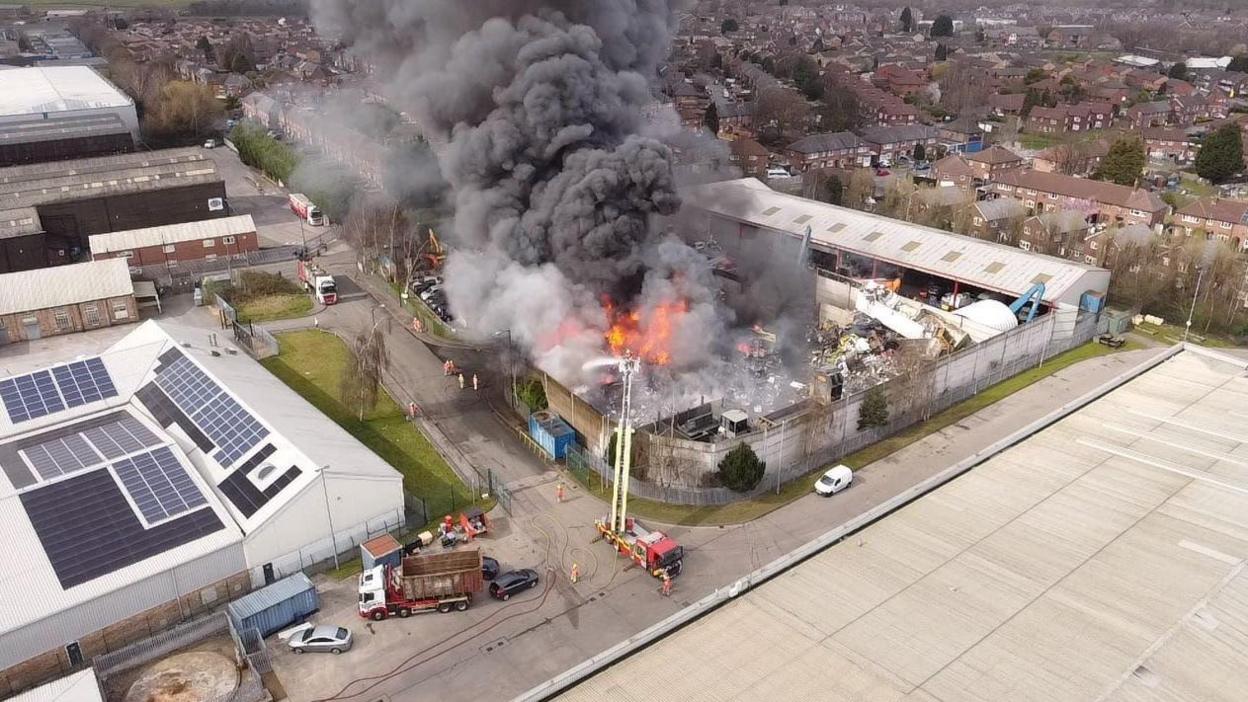 Drone shot of orange flames and dark plumes of smoke rising from a scrapyard, with houses in the background. There is a fire engine in the foreground on the road with a fireman on the ladders dousing the fire.