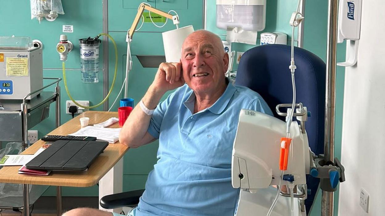 A bald man wearing a blue t-shirt sitting in a hospital chair. There are various bits of medical equipment and machines surrounding him.