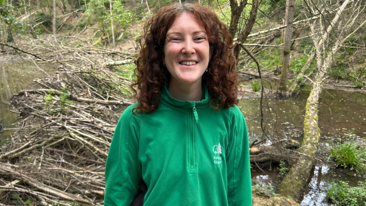 A woman with red curly hair is standing in front of a number of trees in a forest. She is wearing a green jacket with a tree logo and "Forestry England" emblazoned at the front. 