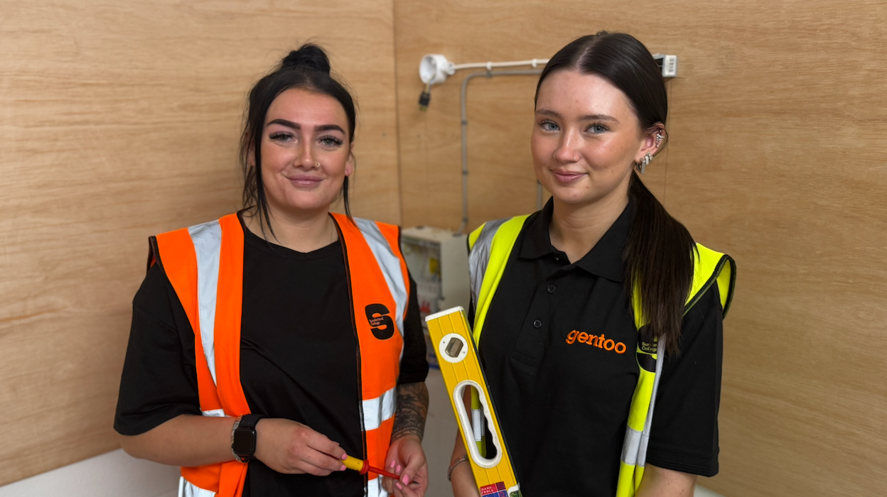 Tammy Cowley, programme lead for electrical installation at Sunderland College, left, is wearing an orange high visibility vest with the Sunderland College logo stands next to one of her students, Emily, who is wearing a black shirt with the orange Gentoo logo printed on the left side. She is holding up a yellow spirit level.