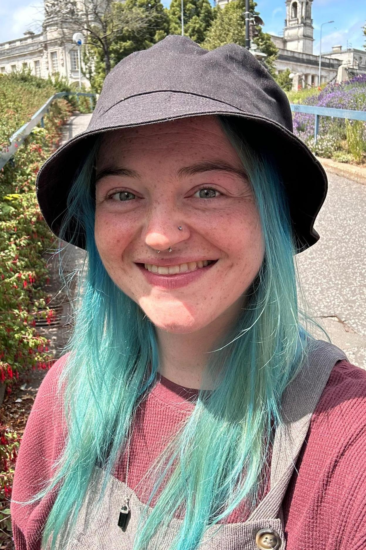 Stephanie has long blue hair is smiling at the camera and wearing a grey bucket hat, pink top and light dungarees. There is a building behind her and bushes to the left. 