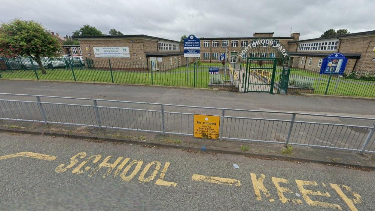 Yellow road markings are on the road in the foreground. The school is behind a fence and the words Northwood Park are above the gate that forms the entrance to the grounds.