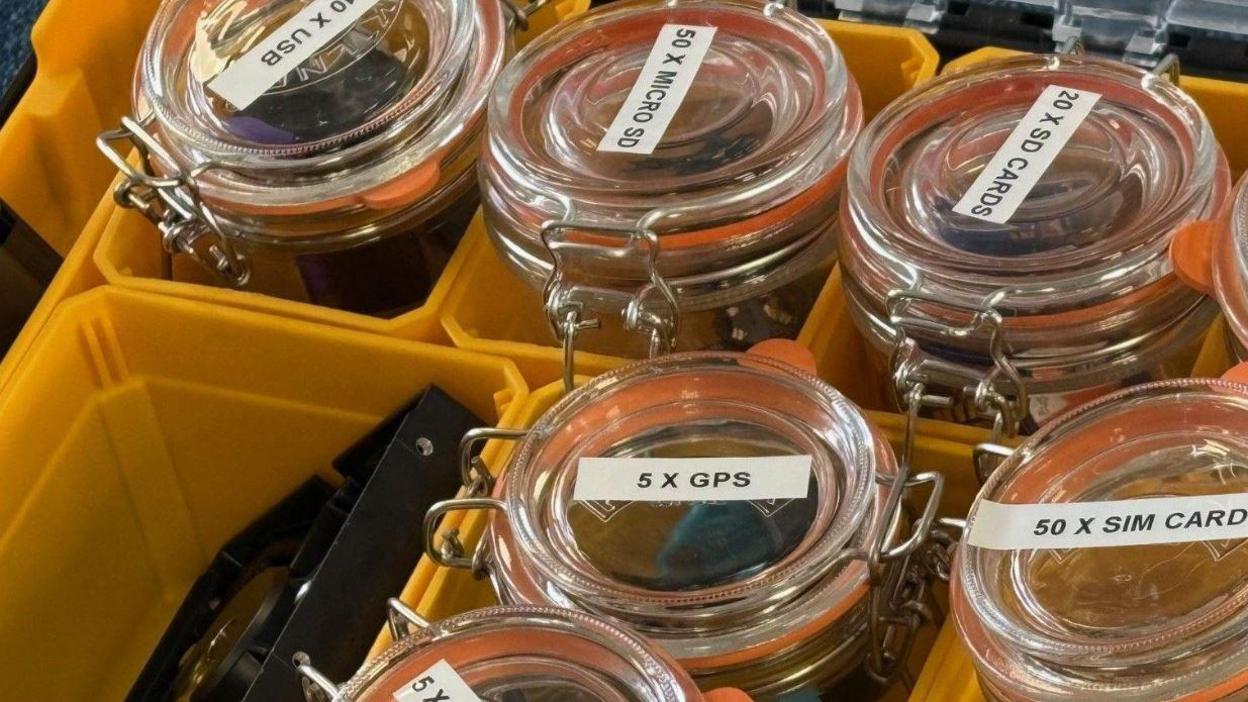 A yellow plastic box divided up into segments. Inside each segment is a glass Kilner jar containing digital items such as USB sticks, mini cameras and SIM cards for mobile phones.