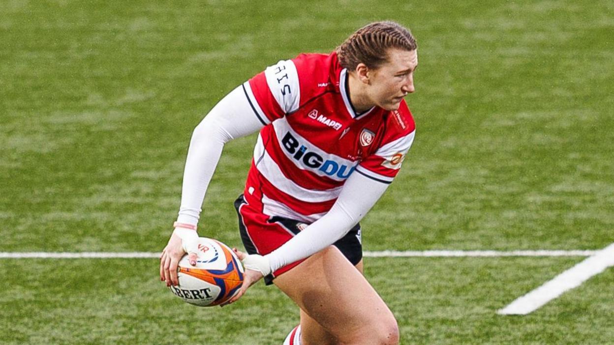 Emma Sing holds the rugby ball down to her right leg in both hands as she looks over her left shoulder while on the pitch during a match for Gloucester-Hartpury