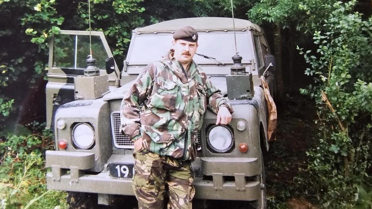 Tony Hutchinson, dressed in camouflage clothing and leaning against an Army Land Rover Defender. Behind the vehicle there's dense green foliage. 