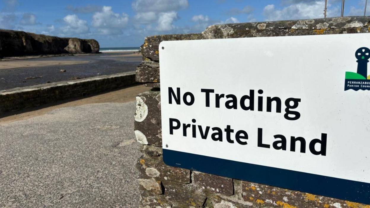 A white sign mounted to a brick wall that reads no trading private land with a beach in the background.