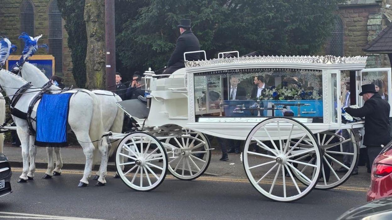 A white horse-drawn carriage, with two white horses pulling it, holds Leo's coffin, which is painted blue with artwork of games consoles on it.