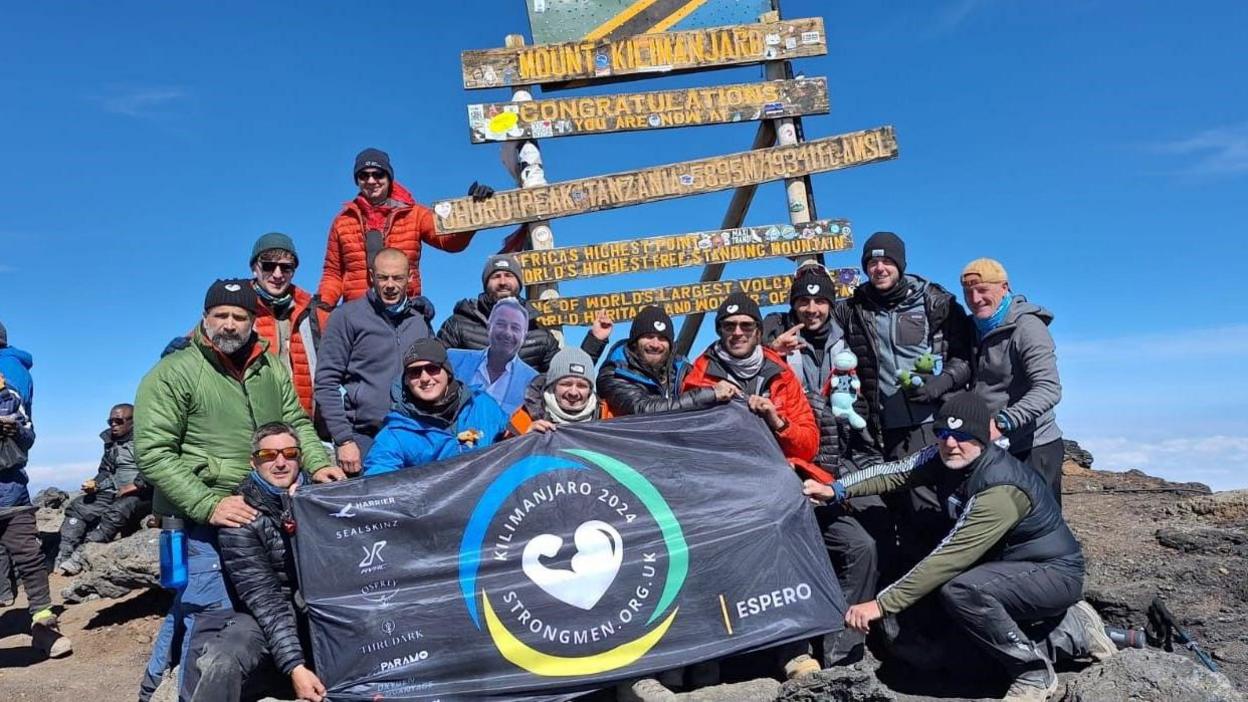 A group of men dressed in mountaineering clothes at the top of Mount Kilimanjaro holding a Strongmen charity banner.