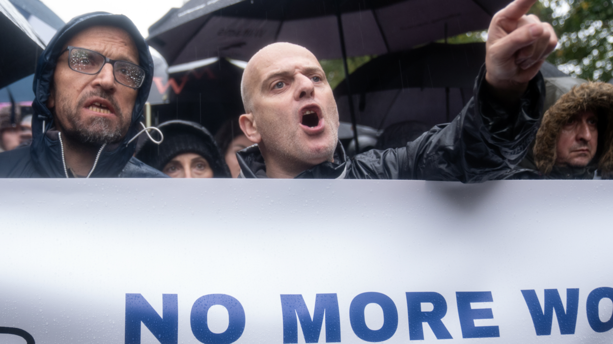 Image shows two men standing behind a banner saying 'NO MORE WORDS'. One is pointing a finger and shouting. 