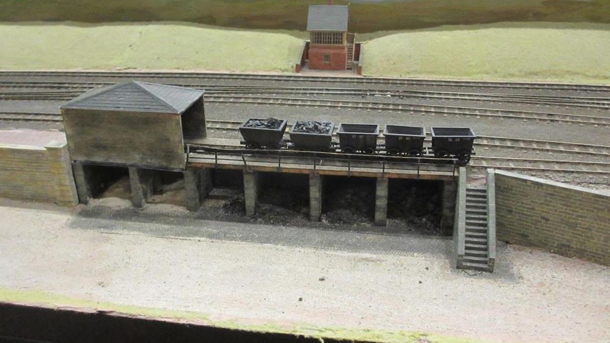 A building is in the foreground with five trailers on the tracks, two filled with coal. There are steps leading down from the track.