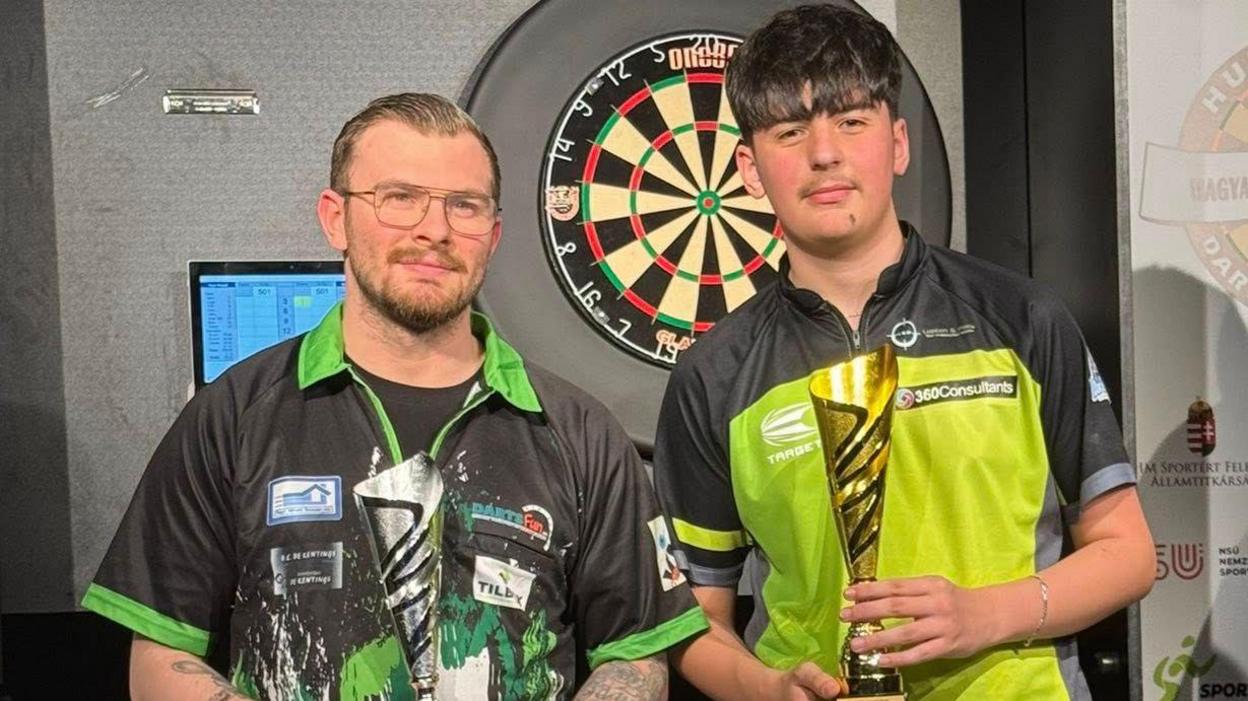 Sybren Gijbels and Kaya Baysal holding trophies after the Budapest Darts Classic final in front of the dart board they played on.