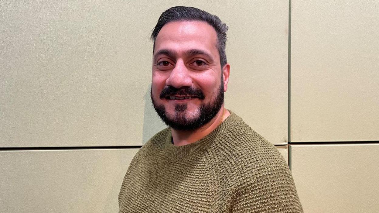 Kulbir wearing a browny-yellow jumper. He is smiling into the camera. He has a beard. He is against a fair coloured backdrop. 